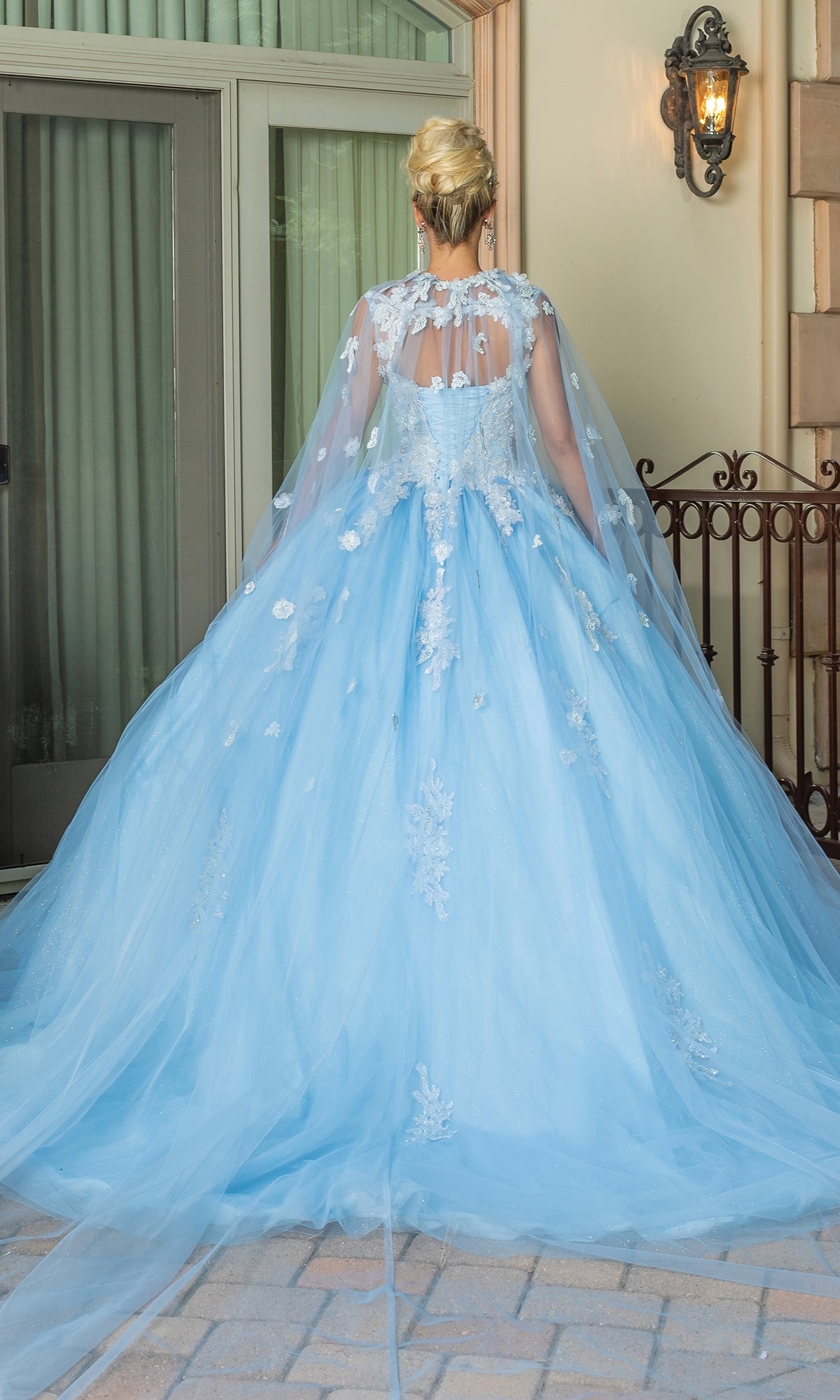 A person with blonde hair in an updo wears the Quinceanera Dress 1597 By Dancing Queen, a light blue ball gown with floral lace embroidery and a long tulle cape, standing outside near a window and wrought iron railing.